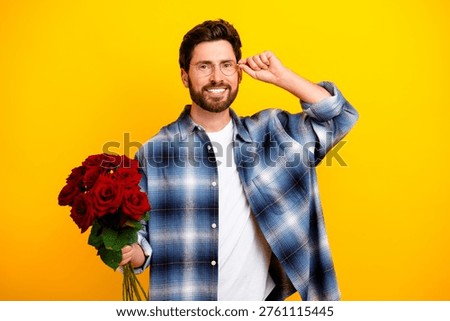 A friendly young man smiles while offering a bouquet of red roses against a bright yellow backdrop, wearing glasses and a casual blue flannel shirt that creates a warm, cheerful romantic mood.