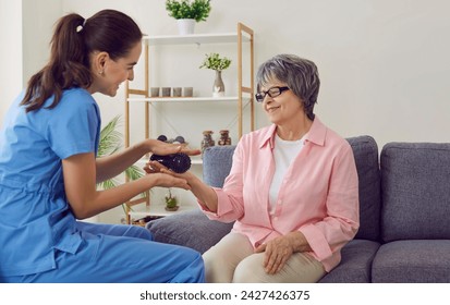 Friendly young nurse or physiotherapist holding spiky rubber ball and doing hand massage to happy smiling senior woman who is sitting on sofa in retirement home. Old age, health, physiotherapy concept - Powered by Shutterstock