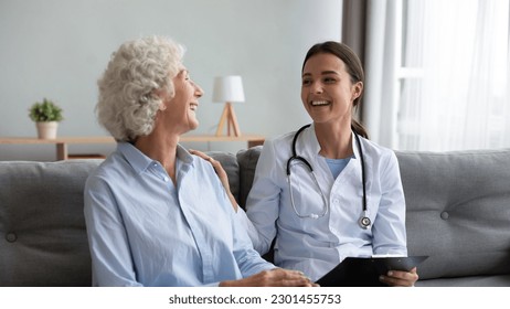 Friendly young nurse in white coat holding clipboard laughing with elderly female patient during homecare visit consultation, provide her support encourages aged woman enjoy warm talk seated on couch - Powered by Shutterstock