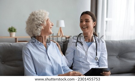 Friendly young nurse in white coat holding clipboard laughing with elderly female patient during homecare visit consultation, provide her support encourages aged woman enjoy warm talk seated on couch