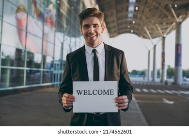 Friendly young satisfied traveler businessman man 20s in black suit stand outside at international airport terminal hold card sign with welcome title text waving hand Air flight business trip concept. - Powered by Shutterstock