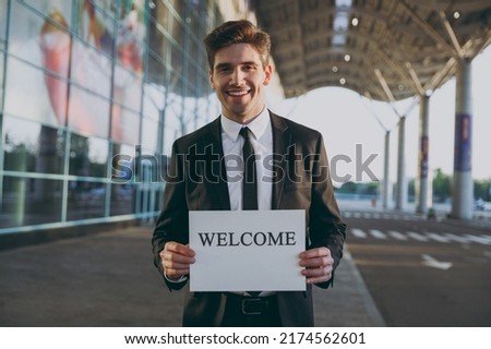 Friendly young satisfied traveler businessman man 20s in black suit stand outside at international airport terminal hold card sign with welcome title text waving hand Air flight business trip concept.