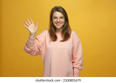 Friendly young smiling girl waving hand, making hello hi welcome gesture, get acquainted on yellow studio background - Shutterstock ID 2100801679