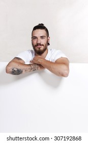 Friendly Young Tattoo Man With Pony Tail Leaning Across A White Wall