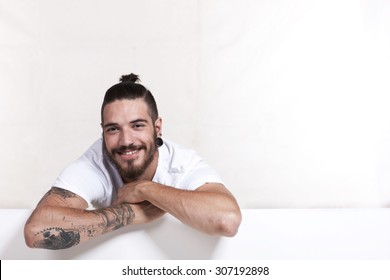 Friendly Young Tattoo Man With Pony Tail Leaning Across A White Wall