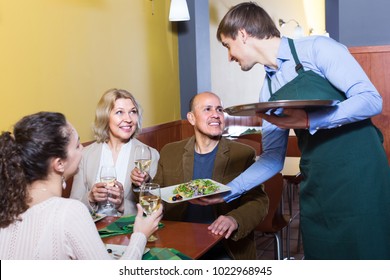 Friendly  young waiter serving table with happy adults in restaurant 