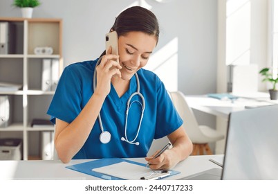 Friendly young woman doctor, nurse, reception specialist in blue scrubs with stethoscope sitting in office workplace, making phone calls, talking to customers, scheduling appointments and taking notes - Powered by Shutterstock