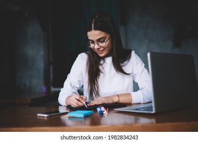 Friendly youthful lady in eyeglasses while sitting at wooden desk in dark workspace with stickers and gadgets while writing down details