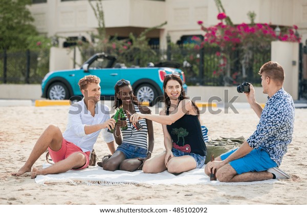 Friends and beer. Four young\
cheerful people taking photo and  cheering with beer and smiling\
while sitting on the blanket on a beach in front of a car and \
villa.