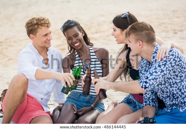 Friends and beer. Four\
young cheerful people cheering with beer and smiling while sitting\
on the blanket.