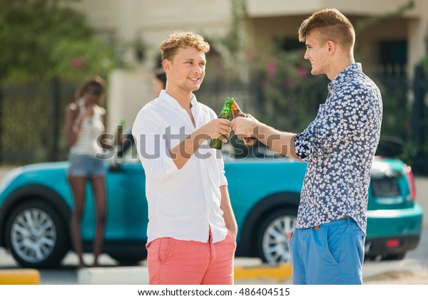 Friends and beer. Four young cheerful\
guys cheering with beer and smiling while staying on a beach in\
front of a car and villa. Boys on a beach drink\
beer.