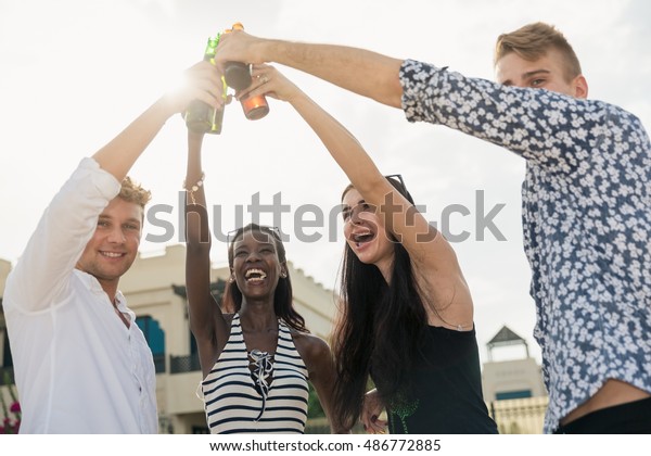 Friends and beer. Four young cheerful people\
cheering with beer and smiling while staying on a beach in front of\
a villa. Guys drink beer on a\
beach.