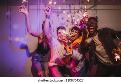 Friends Celebrating And Playing With Fire Sparkles At A House Party. Young Men And Women Having Fun At A Colorful House Party With Decorations And Confetti All Around.