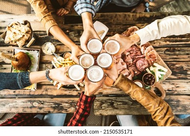 Friends cheering beer glasses on wooden table covered with delicious food - Top view of people having dinner party at bar restaurant - Food and beverage lifestyle concept - Shutterstock ID 2150535171