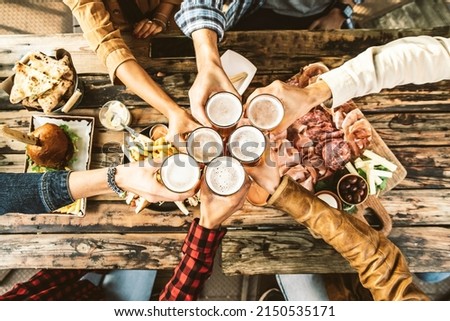 Friends cheering beer glasses on wooden table covered with delicious food - Top view of people having dinner party at bar restaurant - Food and beverage lifestyle concept