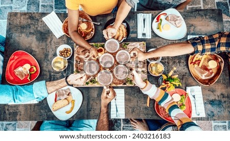 Friends cheering beer glasses on wooden table covered with food - Top view of people having dinner party at bar restaurant - Food and beverage lifestyle concept