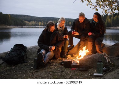 Friends With Coffee Cups Sitting By Campfire On Lakeshore - Powered by Shutterstock