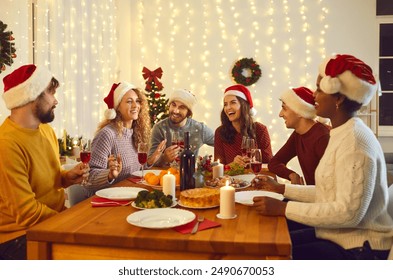 Friends communicate with each other sitting at festive table and enjoying Christmas Eve. Young multiracial people in Santa hats tell funny stories sitting at home in room with Christmas decorations. - Powered by Shutterstock