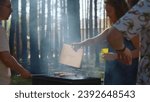 Friends cook meat on coals with grill. Stock footage. Woman blows up coals for grilling meat. Woman waves over grill in woods on sunny day