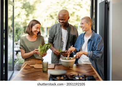 Friends Cooking Salad At Home Kitchen. Black Man And European Girls Enjoying Time Together. Concept Of Healthy Eating. Modern Lifestyle Friendship. Interior Of Apartment. Sunny Daytime