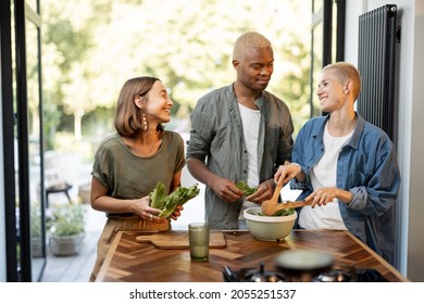 Friends Cooking Salad At Home Kitchen. Black Man And European Girls Enjoying Time Together. Concept Of Healthy Eating. Modern Lifestyle Friendship. Interior Of Apartment. Sunny Daytime