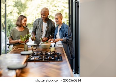 Friends Cooking Salad At Home Kitchen. Black Man And European Girls Enjoying Time Together. Concept Of Healthy Eating. Modern Lifestyle Friendship. Interior Of Apartment. Sunny Daytime