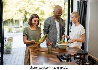 Friends Cooking Salad At Home Kitchen. Black Man And European Girls Enjoying Time Together. Concept Of Healthy Eating. Modern Lifestyle Friendship. Interior Of Apartment. Sunny Daytime