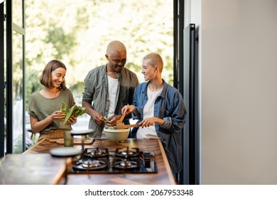 Friends Cooking Salad At Home Kitchen. Black Man And European Girls Enjoying Time Together. Concept Of Healthy Eating. Modern Lifestyle Friendship. Interior Of Apartment. Sunny Daytime