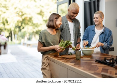 Friends Cooking Salad At Home Kitchen. Black Man And European Girls Enjoying Time Together. Concept Of Healthy Eating. Modern Lifestyle Friendship. Interior Of Apartment