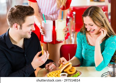 Friends Or Couple Eating Fast Food And Drinking Milk Shakes On Bar In American Fast Food Diner, The Waitress Wearing A Short Costume