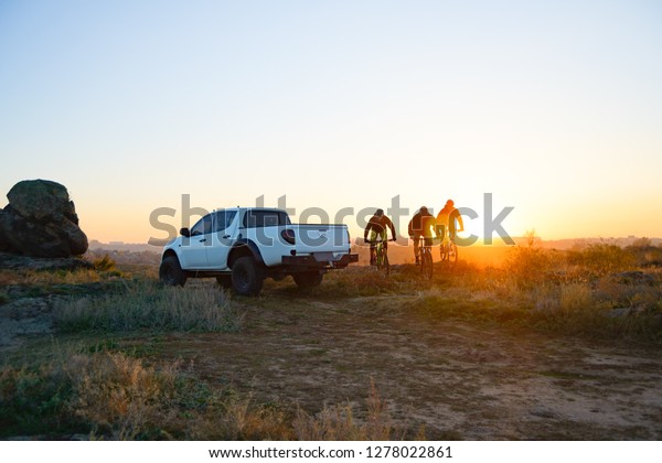Friends Cyclists Riding Enduro Bikes in\
the Mountains in front of the Pickup Off Road Truck at Warm Autumn\
Sunset. MTB Adventure and Car Travel\
Concept.