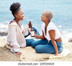 Friends Drinking A Beer On The Beach While Talking And Relaxing On A Summer Tropical Holiday. Happy Black Women Having Fun While On An Ocean Picnic With An Alcohol Drink Enjoying Their Vacation.