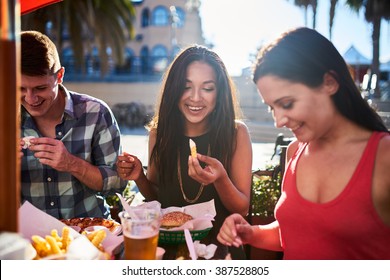 Friends Eating French Fries And Hamburgers Together At Outdoor Restaurant Under Summer Sun