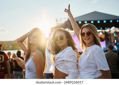 Friends enjoy sunset at beach music fest. Girls dance, laugh in summer vibe. Group celebrates at coastal event with crowd, party atmosphere. Happy young people at outdoor concert, having fun. - Powered by Shutterstock