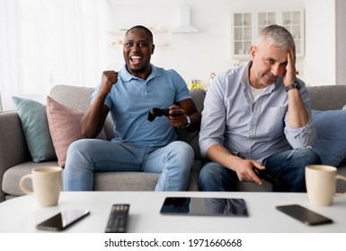 Friends Enjoying Playing Video Game On Playstation, Resting At Home. Sad Elderly Caucasian Man Lose, Happy Mature African American Male Rejoices To Victory With Joystick In Living Room Interior