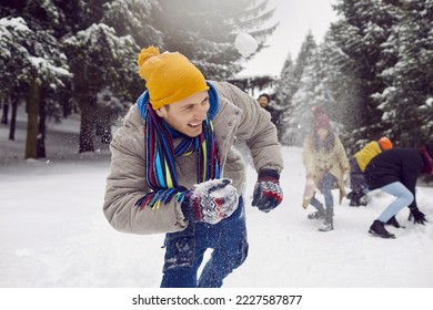 Friends enjoying snowball fight. Young cheerful man laughing out loud while playing snowballs with friends in snowy forest. Funny young man in warm winter clothes dodging snowball. Winter fun concept.