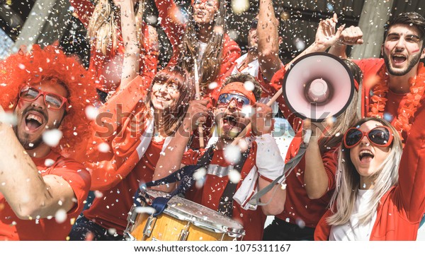 Friends football supporter fans cheering with\
confetti watching soccer match event at stadium - Young people\
group with red t-shirts having excited fun on sport world\
championship concept