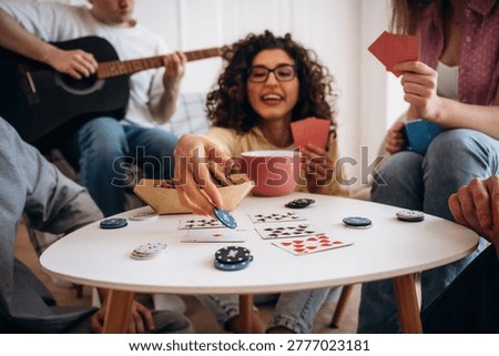 Friends gather around a small table playing cards and sharing snacks, with one person playing guitar in a bright, inviting indoor space