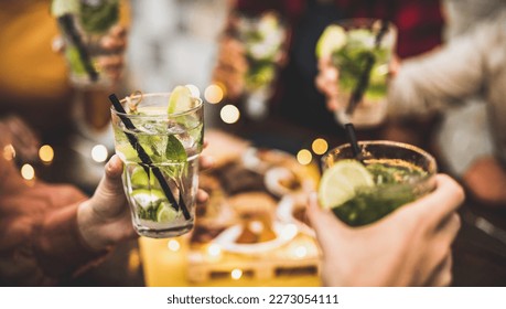 Friends hands toasting mojito drinks at fancy cocktail pub speakeasy - Food and beverage life style concept with people having fun cheering on happy hour at bar - Warm filter with focus on left glass - Powered by Shutterstock