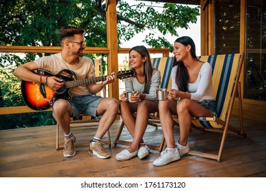 Friends hanging out on vacation at wooden cabin porch, playing guitar and drinking coffee. - Powered by Shutterstock