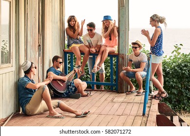 Friends hanging out on vacation at an old wooden cabin porch by the sea while one of them is playing guitar and others are giving him a round of applause - Powered by Shutterstock