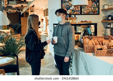 Friends having a chat, holding coffee cups, standing in a coffee shop and waiting for the orders. People wearing the face masks to avoid virus infection and to prevent the spread of disease