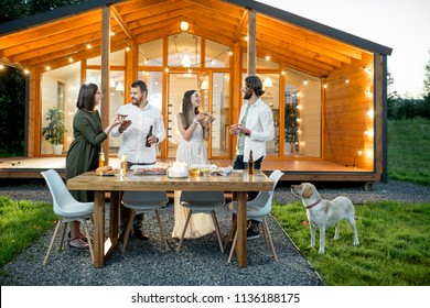 Friends Having A Dinner With Pizza On The Backyard Of The Modern House Decorated With Lights During The Evening Time Outdoors