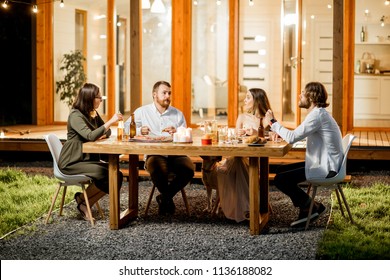 Friends Having A Dinner Sitting At The Table On The Backyard Of The Modern House Decorated With Lights During The Evening Time Outdoors