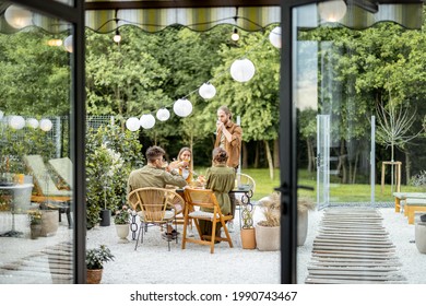Friends having a festive dinner, gathering together at backyard of the house in nature. View through the window from the inside