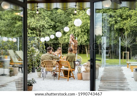 Friends having a festive dinner, gathering together at backyard of the house in nature. View through the window from the inside