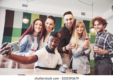 Friends having fun at restaurant. Three boys and three girls making selfie and laughing. On foreground boy holding smart phone. All wear casual clothes.
