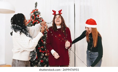 Friends are having fun while decorating a Christmas tree with garlands and lights. They are dressed festively and enjoy each other's company during the holiday season.