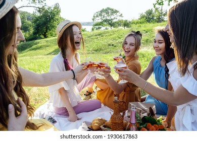 Friends Having Picnic In The Countryside. Group Of Young Women Sitting On Blanket In Park Near Trees, At Sunset On Spring Summer Day. Five Girlfriends Eating And Drinking Wine On Outdoor Party