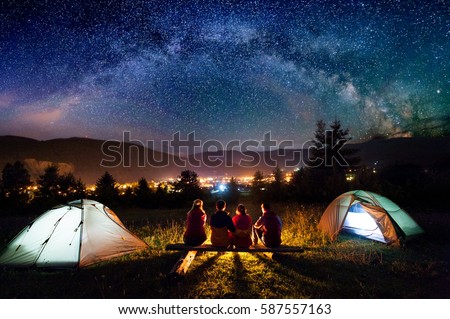 Friends hikers sitting on a bench made of logs and watching fire together beside camp and tents in the night. On the background beautiful starry sky, mountains and luminous town. Rear view